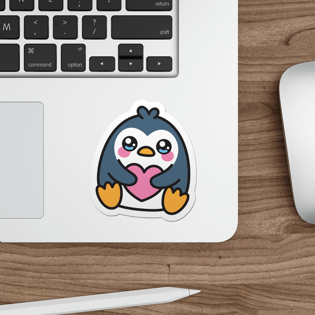 Laptop with a die-cut vinyl sticker of a cute penguin holding a heart on a wooden desk.
