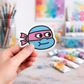 Hand holding a colorful laptop sticker of a cartoon ninja turtle character