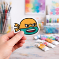 Hand holding a colorful laptop sticker of a cartoon ninja turtle character