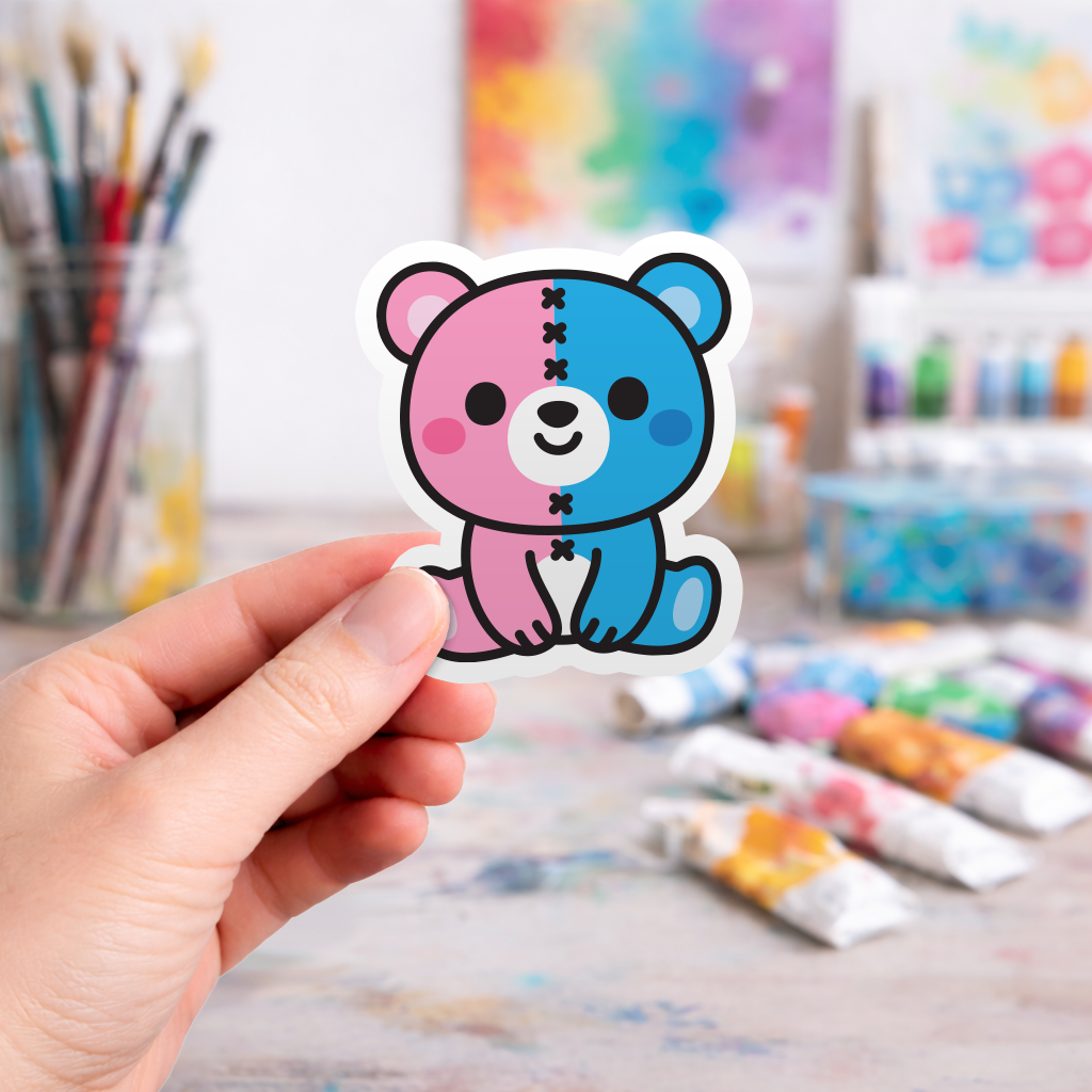 Hand holding a pink and blue bear sticker with craft supplies in the background